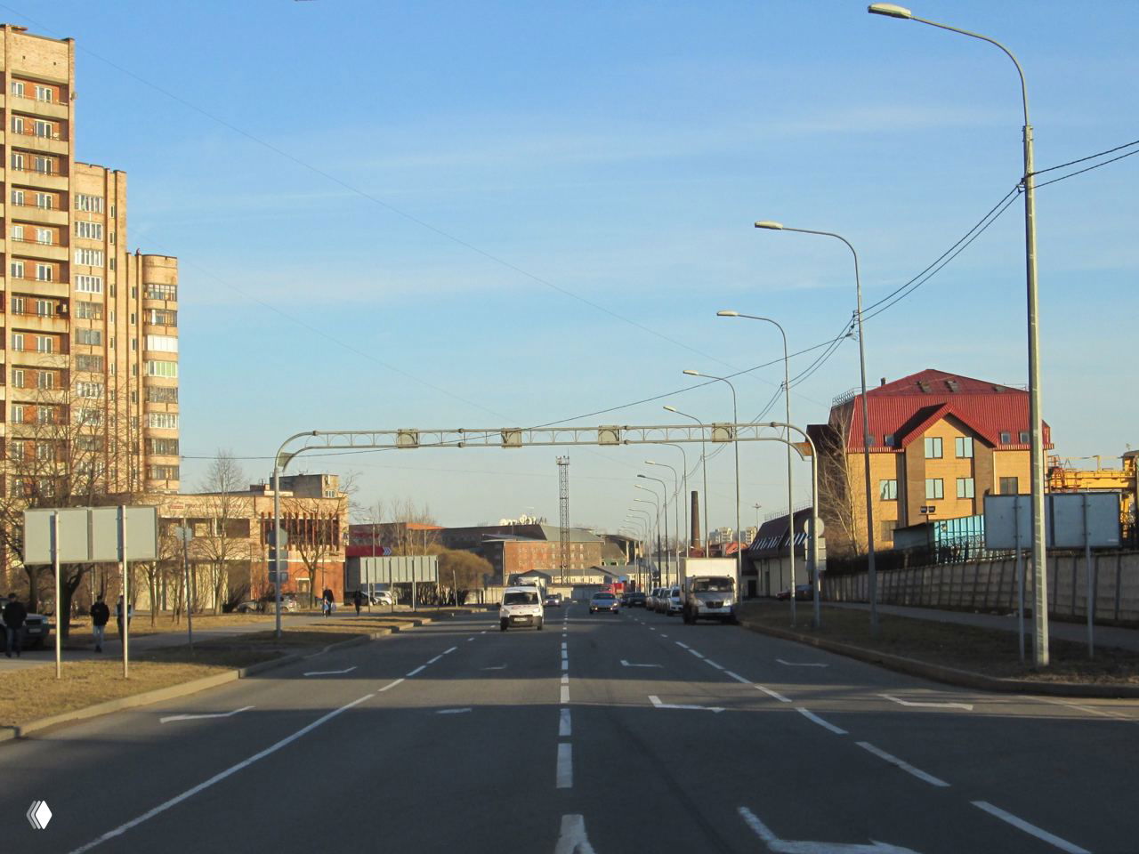 Широкая многополосная улица Петербурга с рядами фонарных столбов, жилыми домами по сторонам и видимой вдалеке промышленной застройкой.