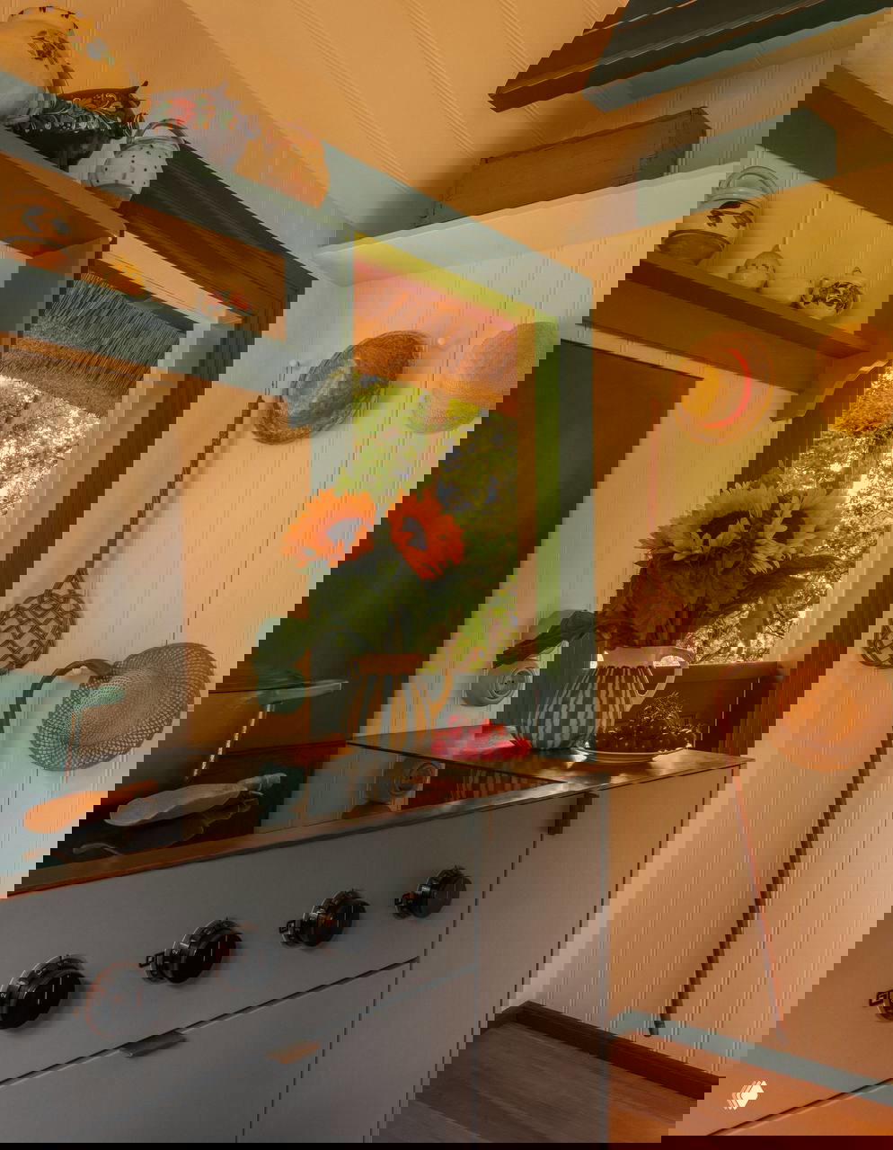 Уголок кухни с синими шкафами, подсвеченными окнами и букетом подсолнухов в кувшине на подоконнике; на стене висят соломенные шляпы и кухонные аксессуары.