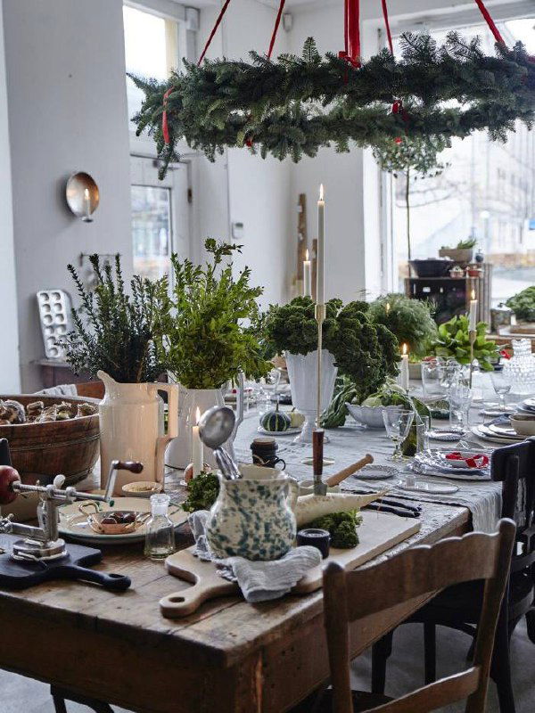 Круглая подвесная композиция из еловых ветвей над столом, свечи и зелёные акценты создают естественный зимний декор вместо привычной елки.