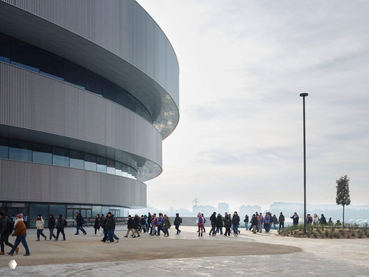 Вид фасада Santagiulia со стороны входной площади днём: изогнутые металлические полосы, остеклённые секции и люди у входа.