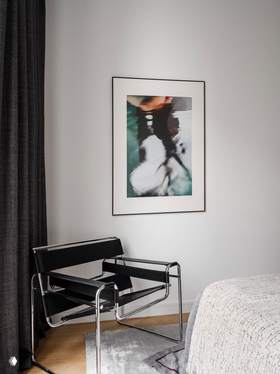 Черное кресло-«вассерман» с хромированной рамой в углу комнаты и абстрактная фотография в раме на светлой стене.