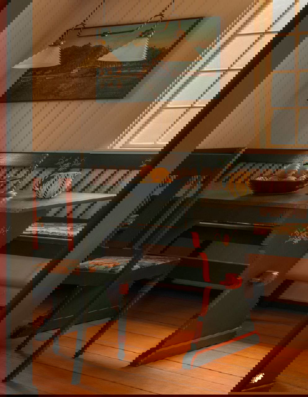 Обеденная зона с резным деревянным столом и скамьёй в тёмно-зелёных и красных тонах, окно пропускает мягкий свет, подчёркивающий натуральные текстуры интерьера.