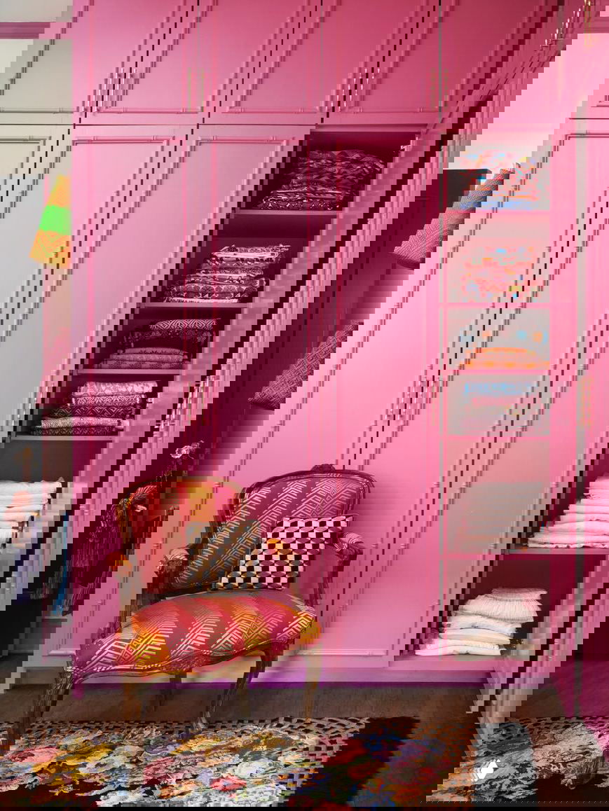 Гардеробная зона с ярко‑розовыми шкафами и антикварным креслом на ковре с цветочным орнаментом, смелый пример работы с цветом в интерьере.