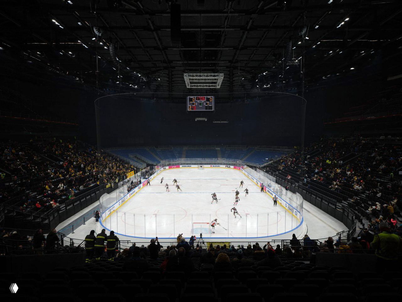 Внутренний вид ледовой площадки Santagiulia: хоккейный лёд, борта и два яруса трибун с сидящими зрителями и табло в центре.