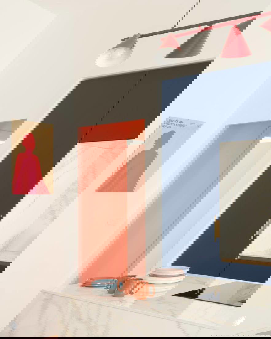 Фрагмент интерьера с ярко-коралловым цветом дверного проёма и синими фасадами кухни, на переднем плане мраморная поверхность и посуда.