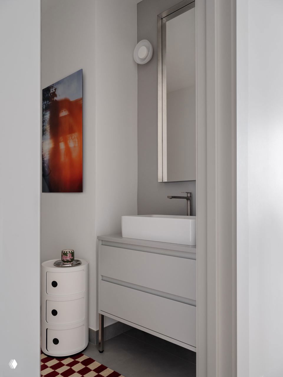Мини-санузел с компактной тумбой и раковиной, светлая лаконичная мебель и небольшая яркая картина у входа.