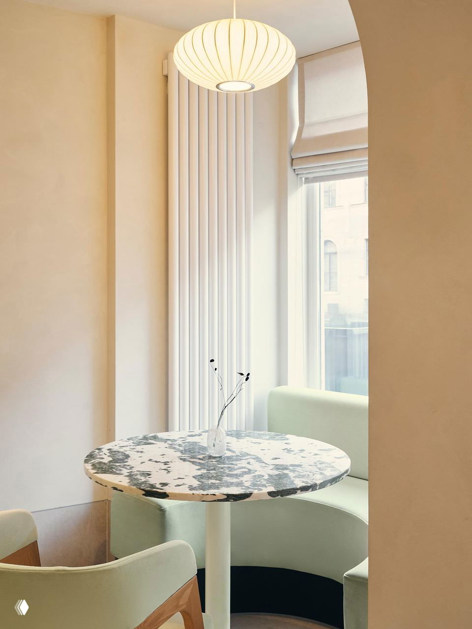 Уголок у окна с мраморным столом, округлыми мягкими сиденьями и высокими вертикальными радиаторами на фоне светлого фасада — дневной свет подчёркивает текстуры интерьера.