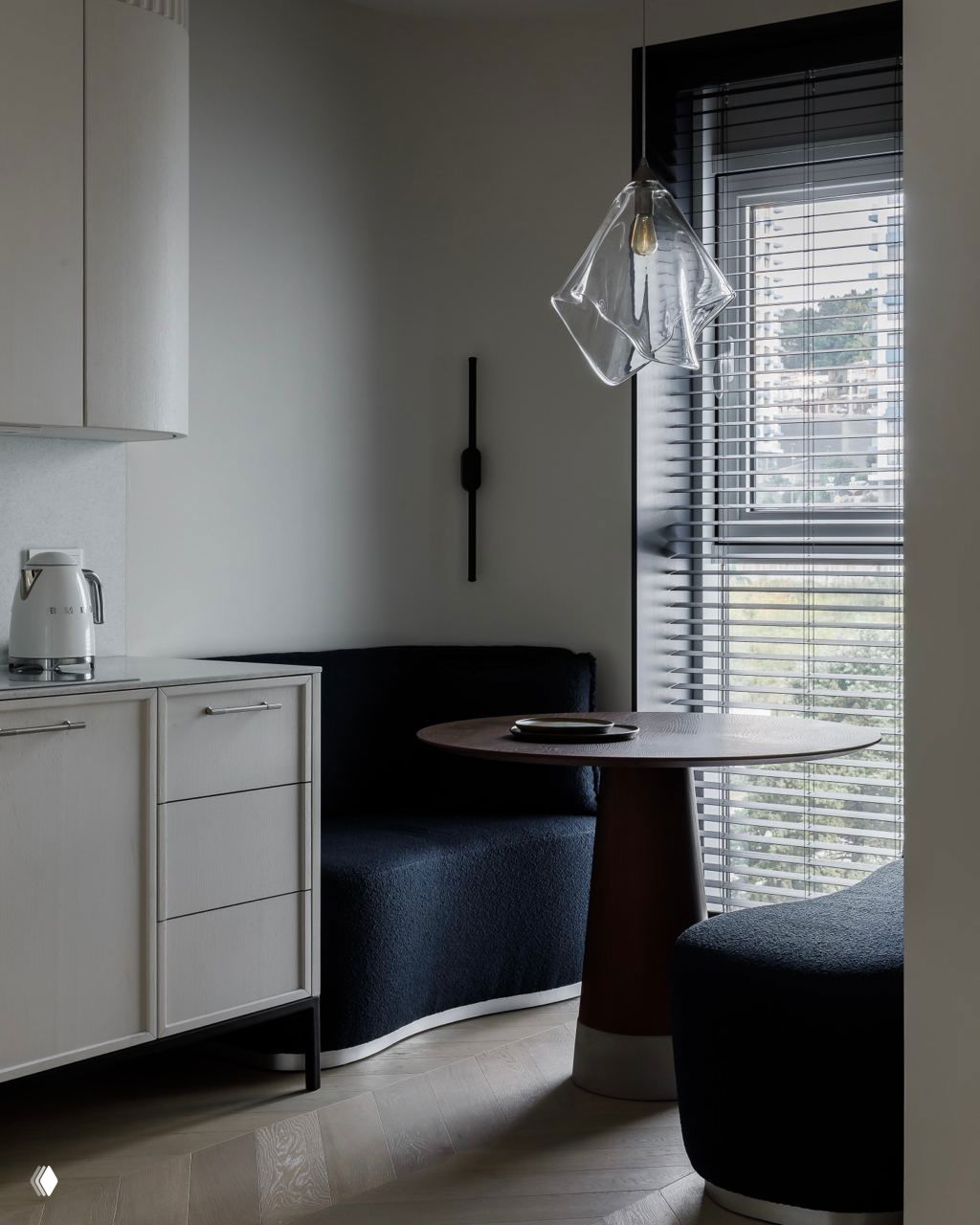 Обеденная зона с круглым столом и тёмным модульным диваном у окна: стеклянная подвеска и жалюзи формируют спокойную атмосферу.