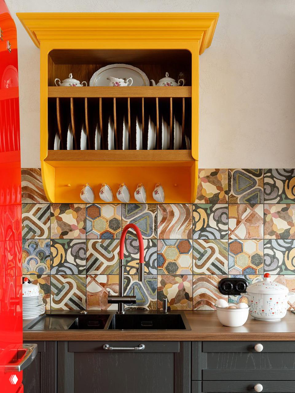 Яркий художественный фартук из разноцветной декоративной плитки с орнаментом, желтая полка для тарелок и красные элементы техники на рабочей стене.