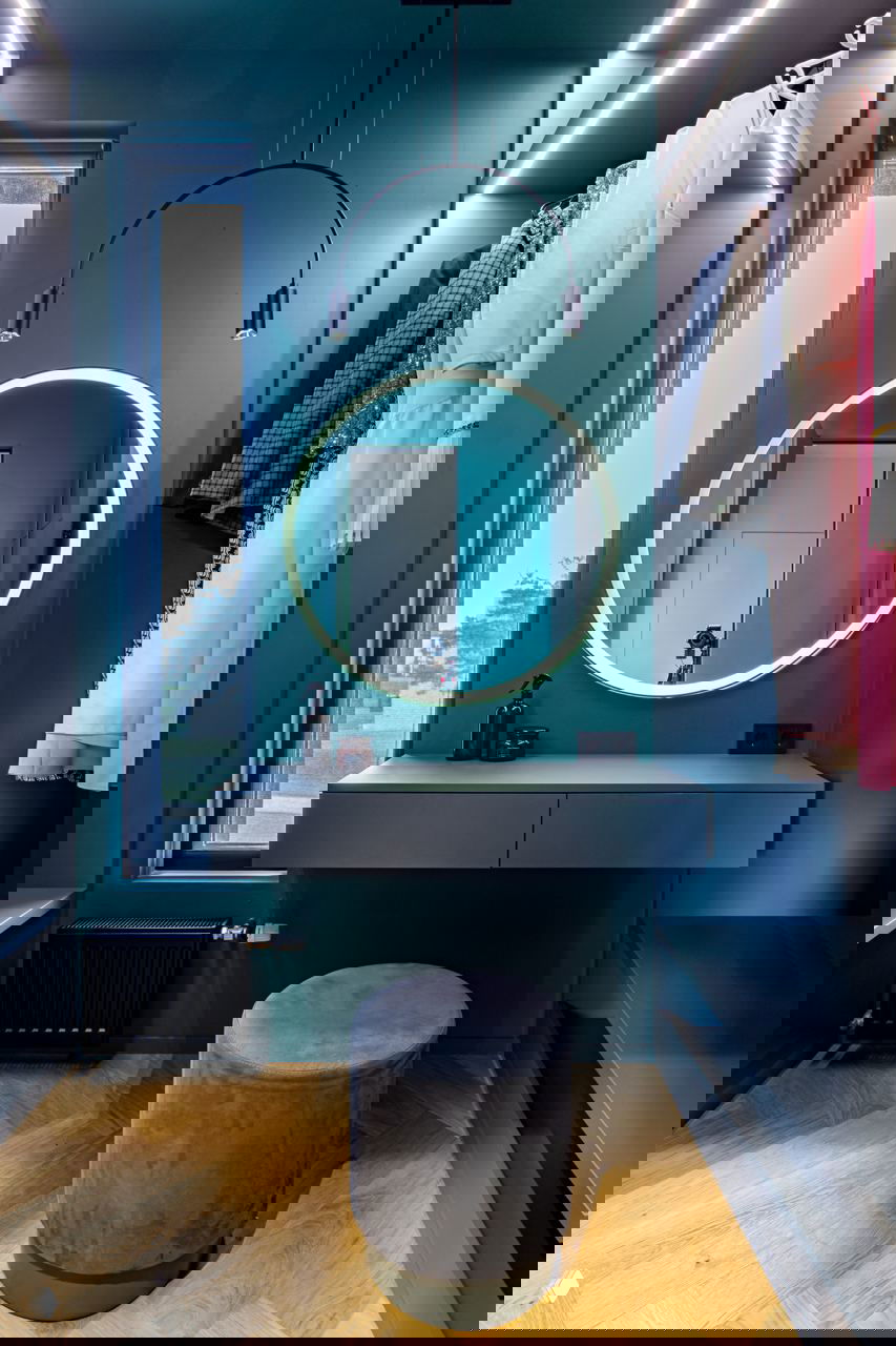 Современная гардеробная в насыщенных тонах с круглым зеркалом с подсветкой, подвесными лампами и небольшим пуфом у туалетного столика.