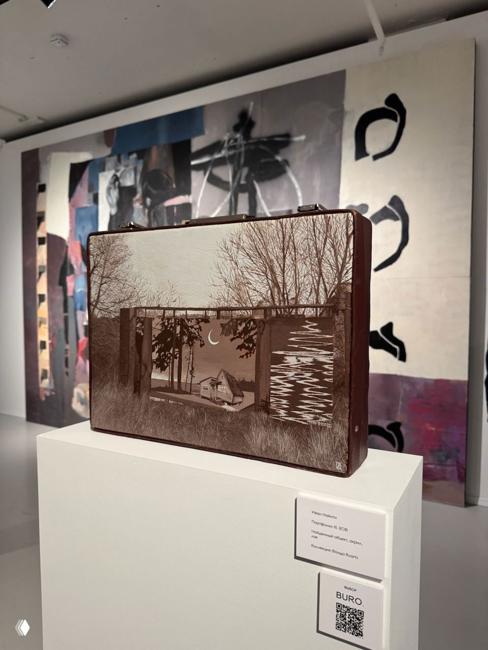 Скульптурная работа в виде чемодана на постаменте на фоне большого полотна; музейная этикетка и логотип проекта рядом.