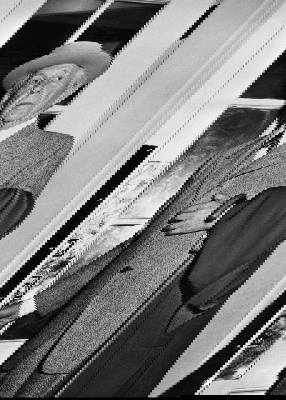 Чёрно‑белый портрет пожилого мужчины в пальто и шляпе, стоящего у двери и опирающегося на трость; формальный интерьерный кадр с выразительной мимикой.