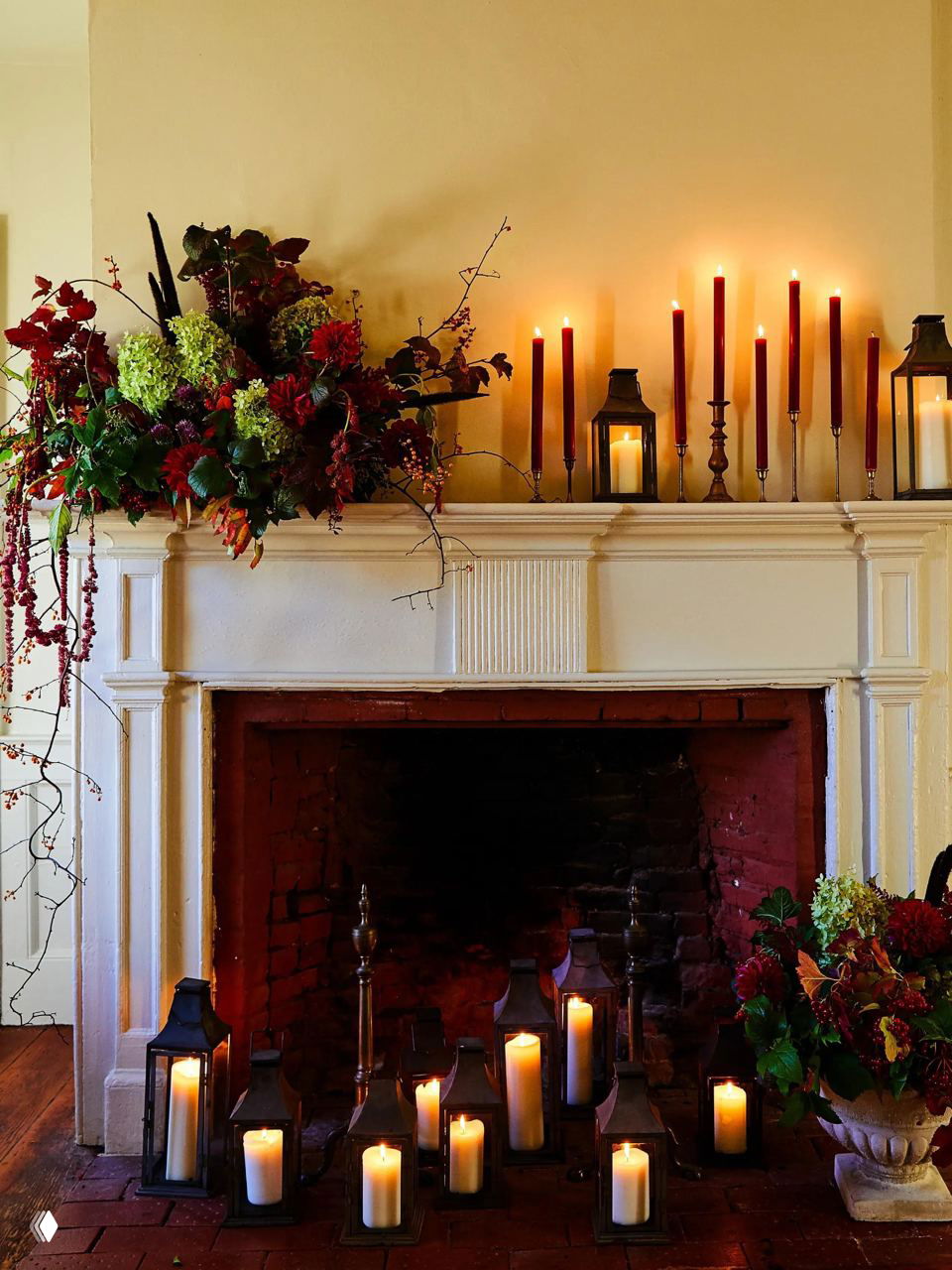 Каминный портал, окруженный множеством фонарей и свечей, с большим цветочным аранжированием и мягким теплым светом.
