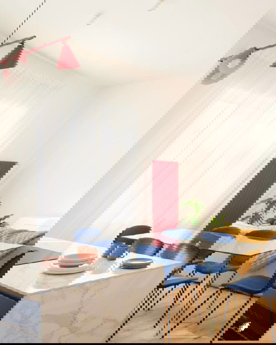 Обеденная зона с прозрачным стеклянным столом, синими стульями, мраморным островом и большими светлыми окнами, мягкая дневная подсветка.