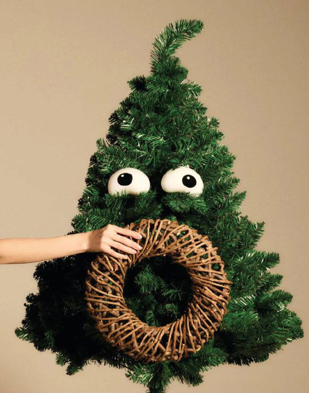 Изображение искусственной елки с большими круглые глазами-игрушками и плетеным венком в руках модели — шарики Treebies от Alessi выглядят как «глаза».