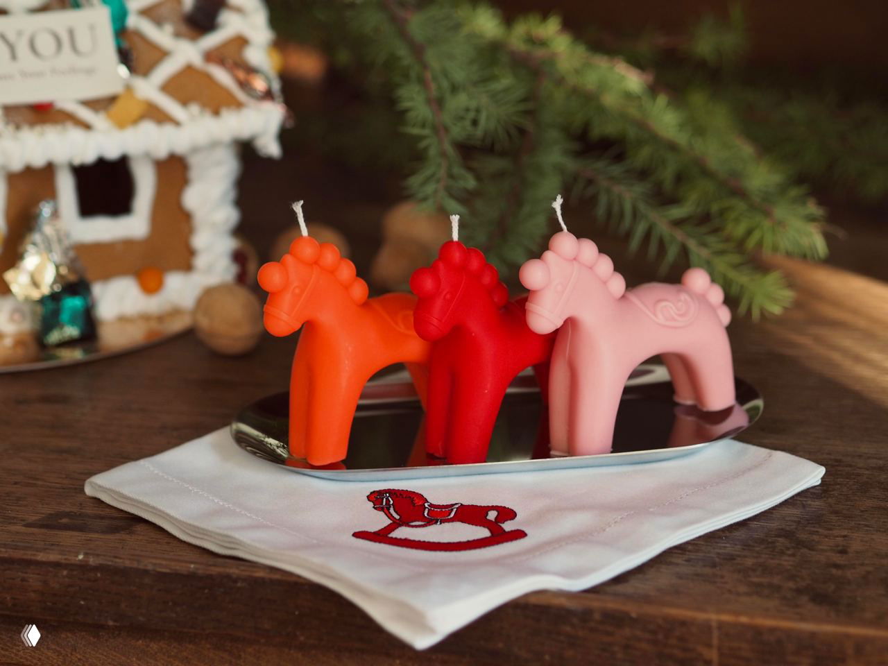 Три восковые свечи в форме лошадок на подносе: оранжевая, красная и розовая, стоящие на салфетке с изображением игрушечной качалки.
