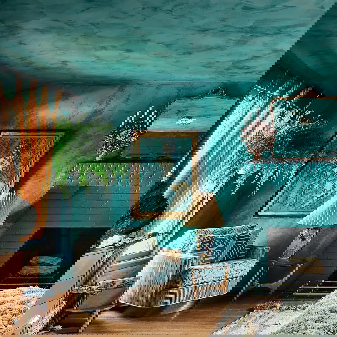 Комната с бирюзово-зелёной декоративной штукатуркой, винтажным креслом у камина и растениями у окна в экологичном интерьере.