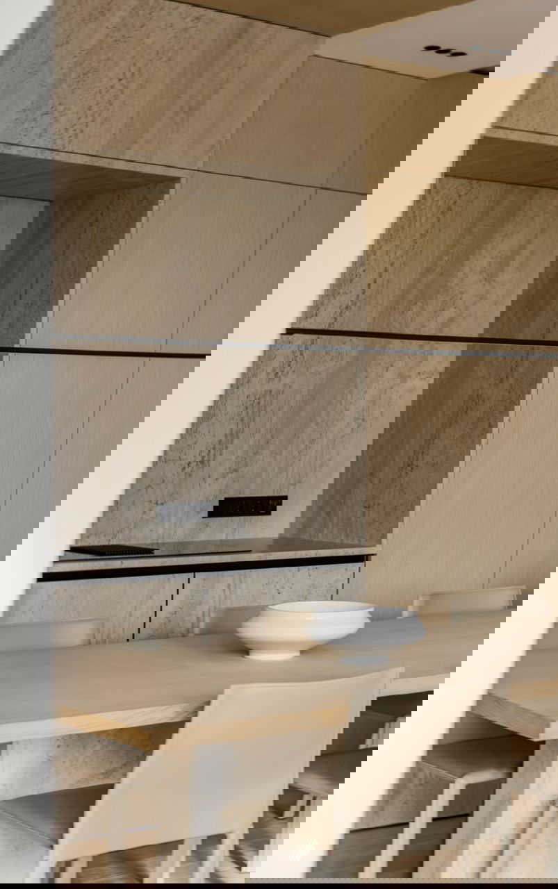 Обеденная зона и кухня: стол из светлого дерева, натуральный камень на фартуке и встроенные тумбы в единой бежевой палитре минималистского интерьера.