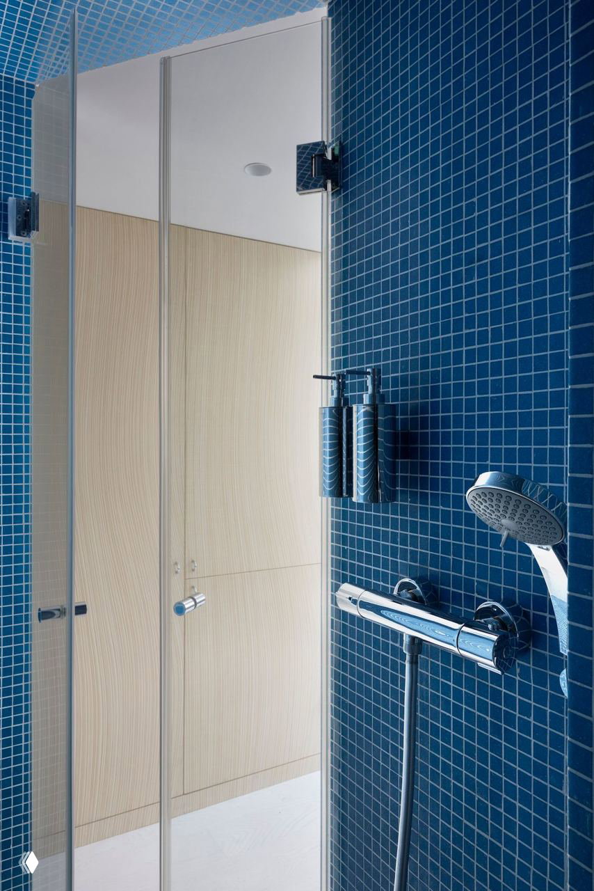 Голубая мозаичная душевая кабина с хромированной сантехникой на фоне светлых деревянных поверхностей — яркий акцент в небольшой квартире.