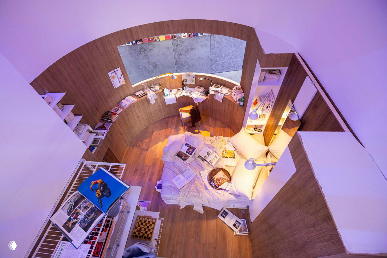 Вид сверху на круглую уютную нишу-инсталляцию: деревянный настил, подушки, разбросанные журналы и постельное место, создающие атмосферу домашнего уюта.