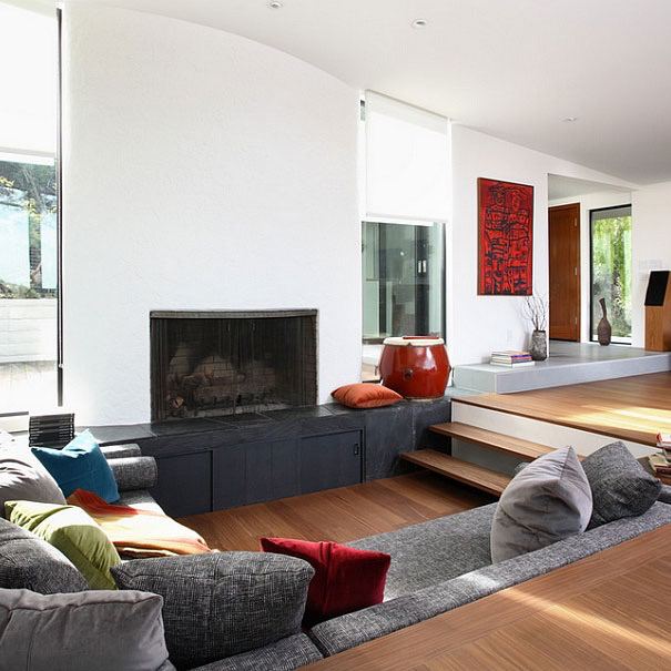 Гостиная с пониженной «утопленной» зоной и ступенчатой планировкой, мягкие подушки и деревянные элементы у камина, светлая композиция.