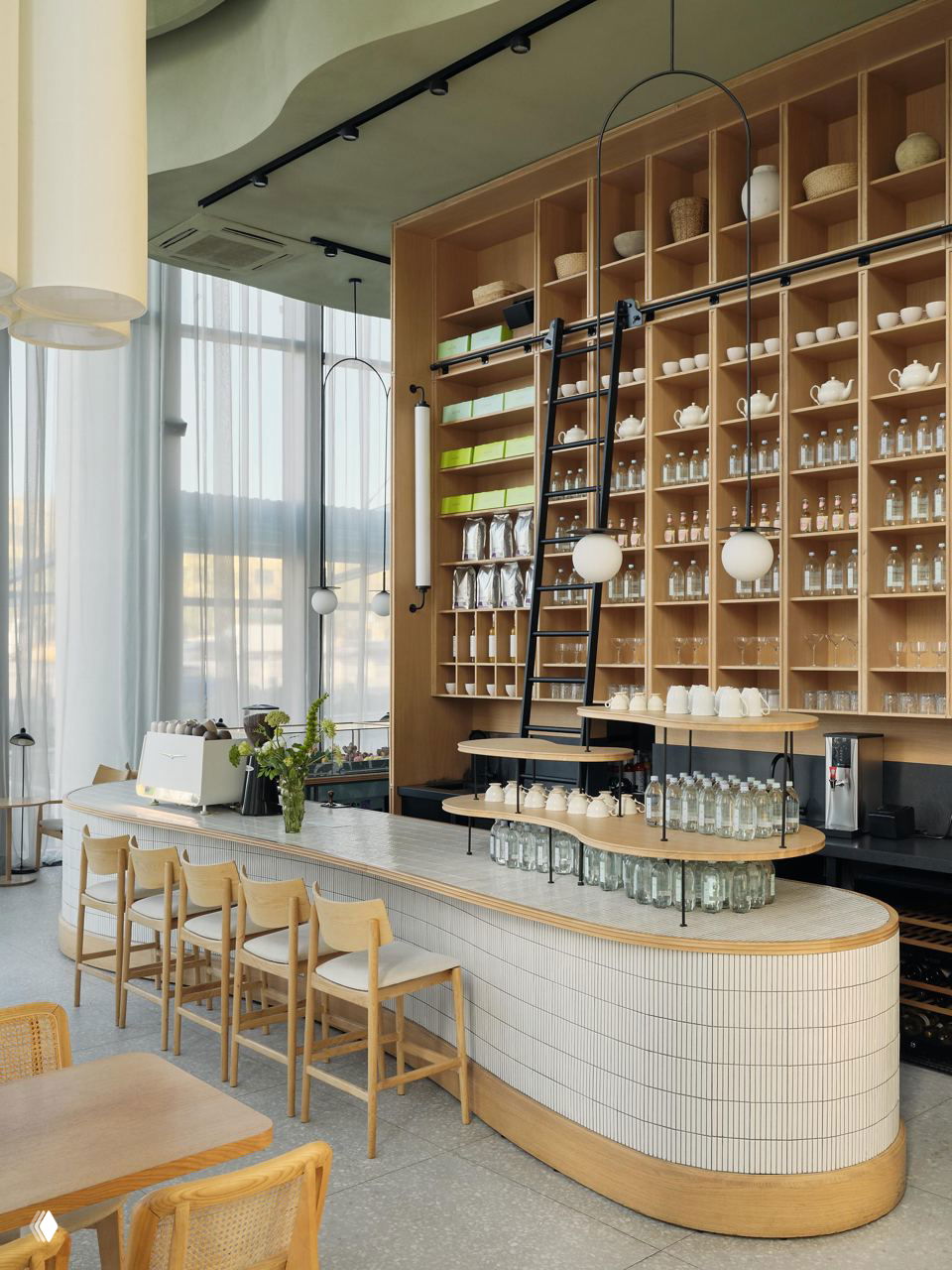 Барная зона с высокой деревянной стеллажной полкой, лестницей для доступа к полкам, белая мозаичная барная стойка и светлая деревянная мебель