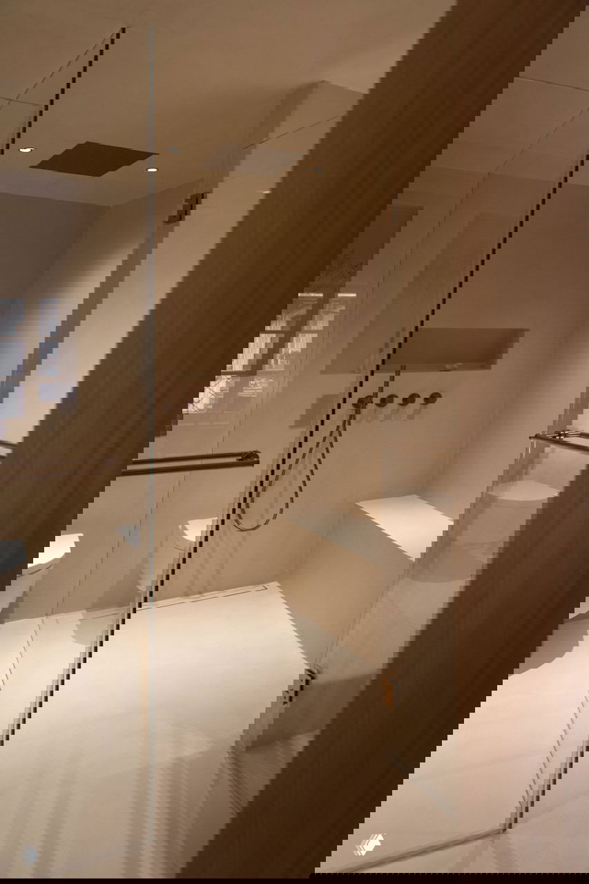 Душевая зона с прозрачной перегородкой, сиденьем и скрытыми смесителями на фоне ровных светлых стен и деревянной двери.