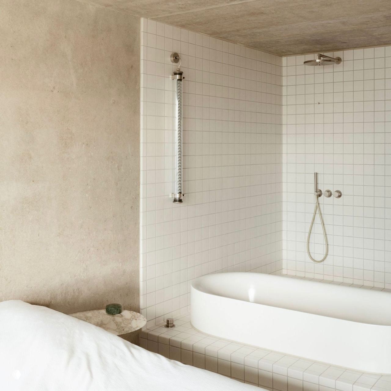 Белая овальная ванна на подиуме и плиточная стена с душем, чистые линии и простая отделка для удобного уборочного ухода в спальнeй зоне.