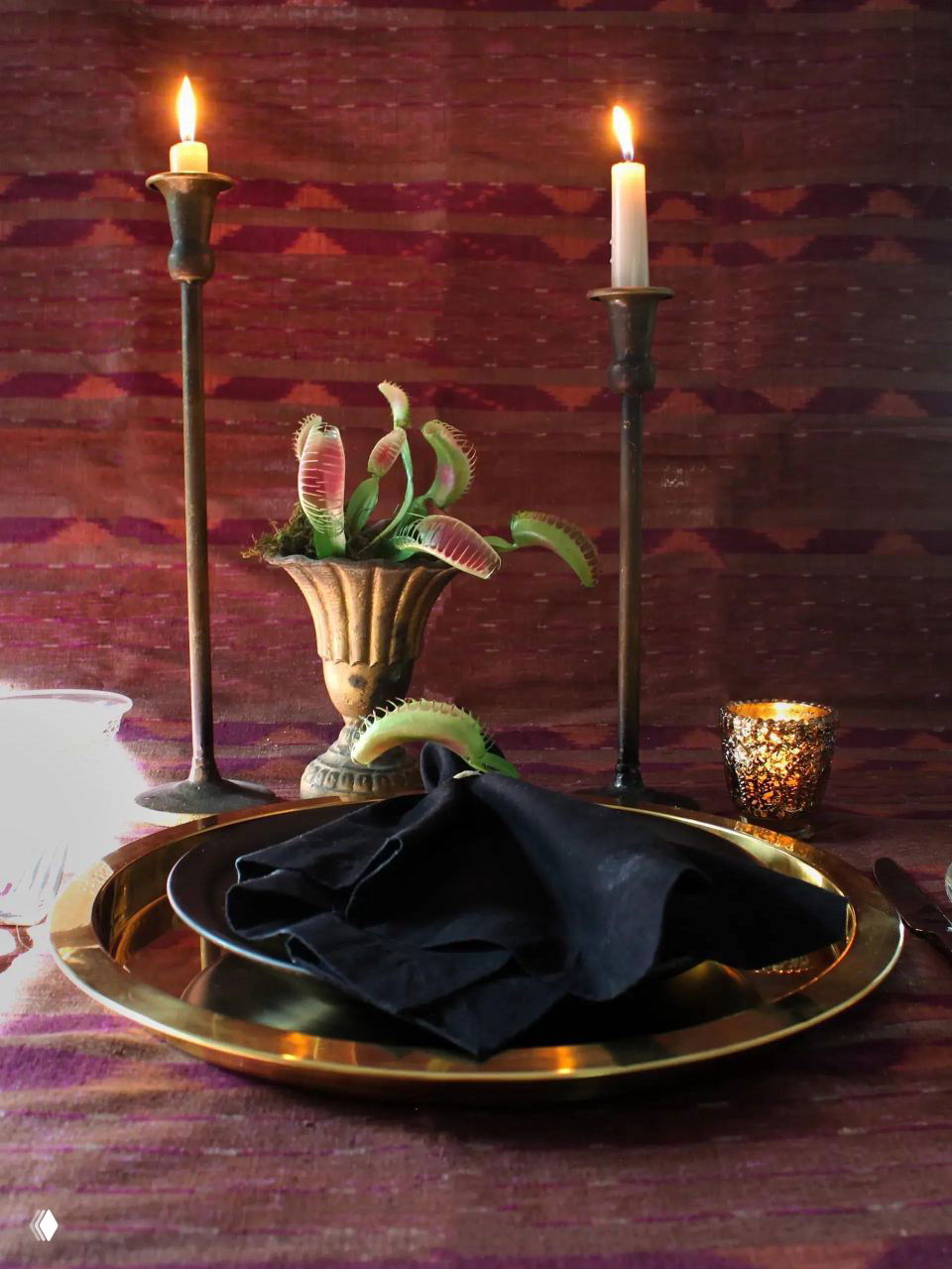 Интимная сервировка с золотой тарелкой, темной тканевой салфеткой и парой свечей на металлических подсвечниках на текстильном фоне.