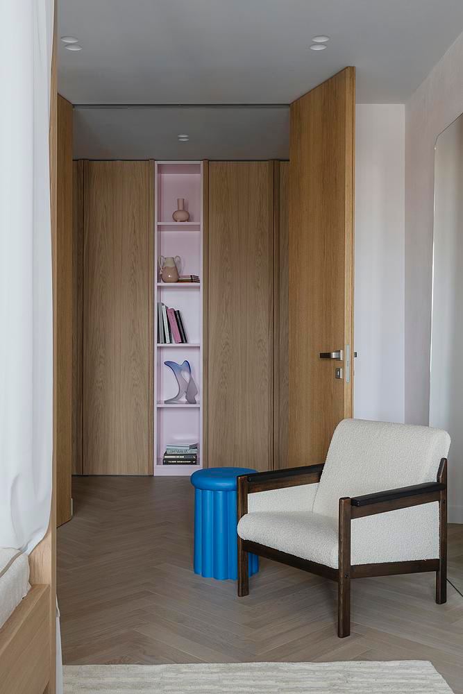 Проход с встроенным деревянным шкафом и центральной нишей, розовая подсветка в нише, синий табурет и кресло в мягкой текстуре — спокойная прихожая в скандинавском духе.