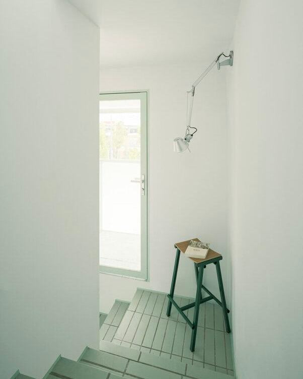Узкая лестничная зона бывшей сторожевой башни с минимальной мебелью: высокий табурет и настенный светильник на фоне белых стен и входной двери.