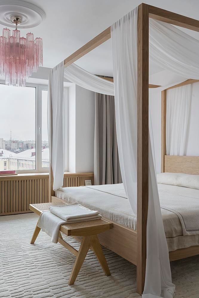 Спальня с четырехстоечной кроватью, легкими белыми драпировками и розовой хрусталистой люстрой над окном; текстиль и дерево создают нежную атмосферу комнаты.