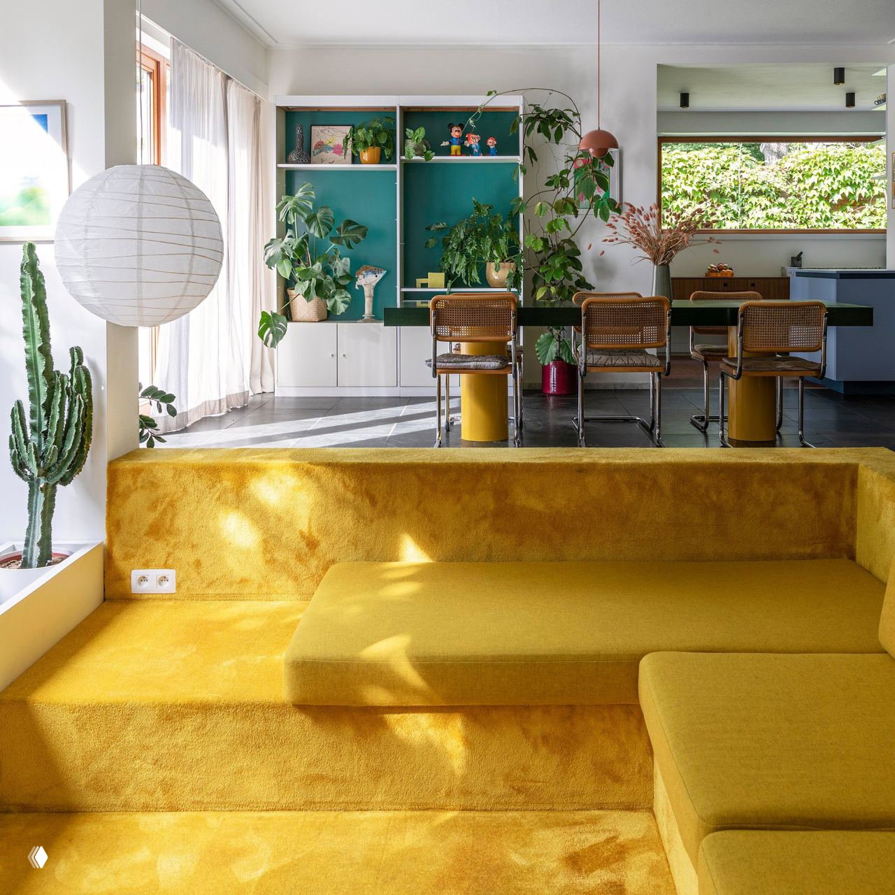 Интерьер с платформенной мягкой системой сидений, обшитой жёлтым текстилем, на фоне столовой с зелёными акцентами и растениями у окна.