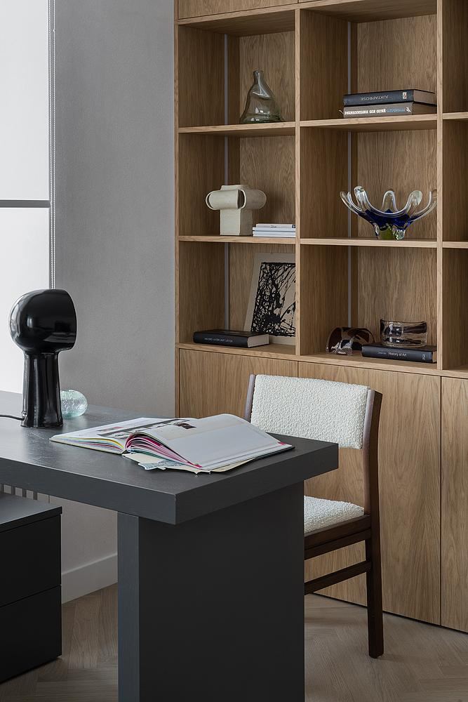 Рабочая зона: темный стол, открытые деревянные стеллажи с книгами и декоративными объектами, столешница с журналом и скульптурной лампой — функциональный уголок в квартире.