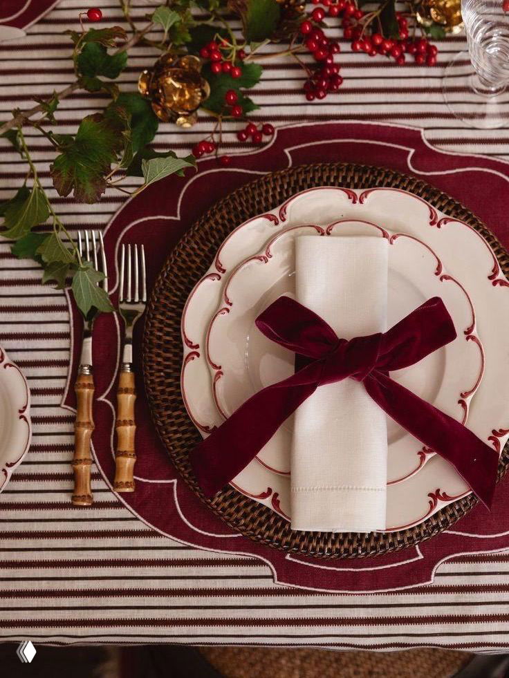 Крупный план тарелки с декоративной каймой, бордовый бант на белой салфетке и текстиль с полосатым узором — элегантная деталь праздничного стола.