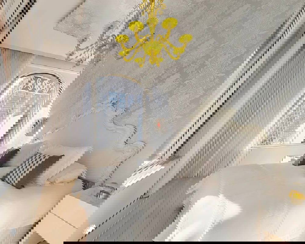 Ещё одна светлая спальня пентхауса с богато декорированным изголовьем, высоким окном и желтой люстрой — сочетание классики и эклектики.