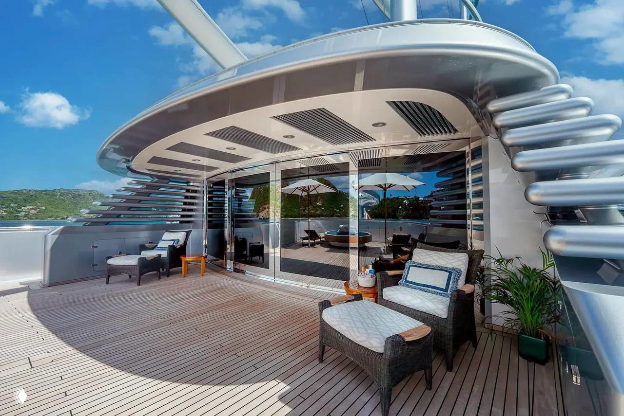 Открытая палуба яхты Maltese Falcon с лаундж-зоной: плетёная мебель, деревянный настил и панорамные раздвижные двери на фоне неба и воды.