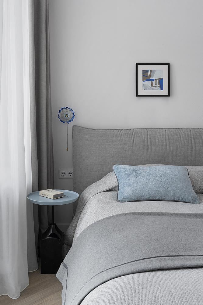 Нейтральный изголовье кровати в светло-серых тонах с декоративной голубой подушкой и небольшим круглым прикроватным столиком — спокойная спальня с акцентом на фактуры.