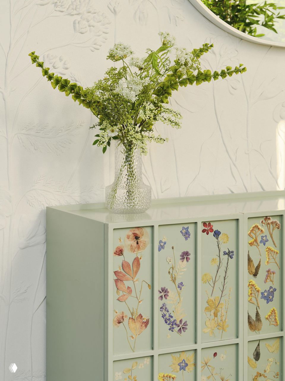 Крупный план цветочной композиции в стеклянной вазе на фоне тиснёной стены и комода с отпечатками засушенных цветов в пастельной гамме