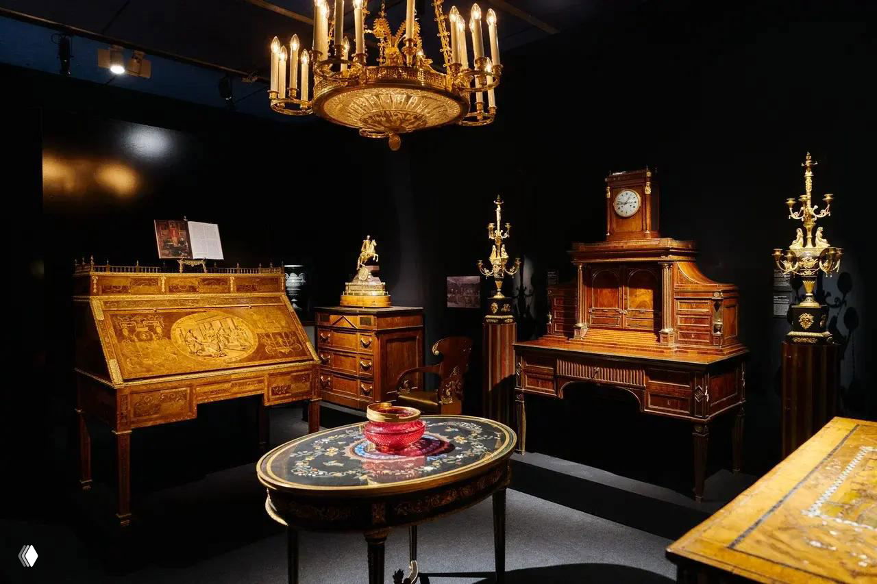 Экспозиция антикварной мебели и декоративных предметов: резные комоды, письменный стол и люстра в тёплом выставочном освещении, классическая обстановка.
