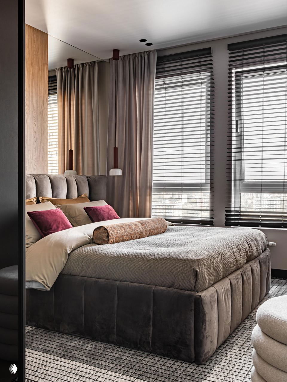 Спальня в тёплой гамме: высокая бархатная кровать, текстильные подушки бордово‑зелёных оттенков и большие окна с горизонтальными жалюзи.