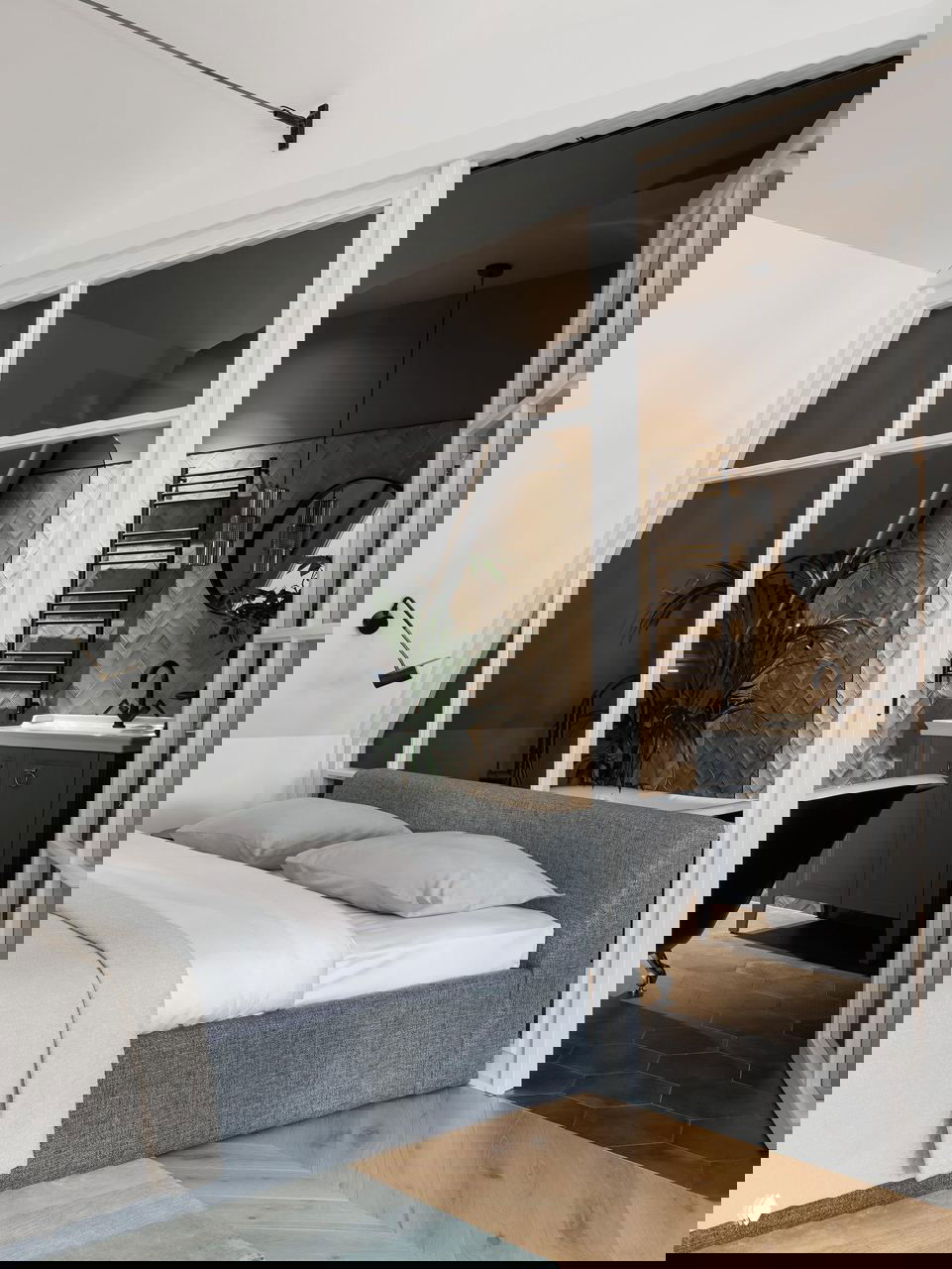 Спальня и ванная, разделённые стеклянной перегородкой: серый диван-кровать, мягкие текстуры и элегантная чёрная ванна на фоне тёплого паркета.