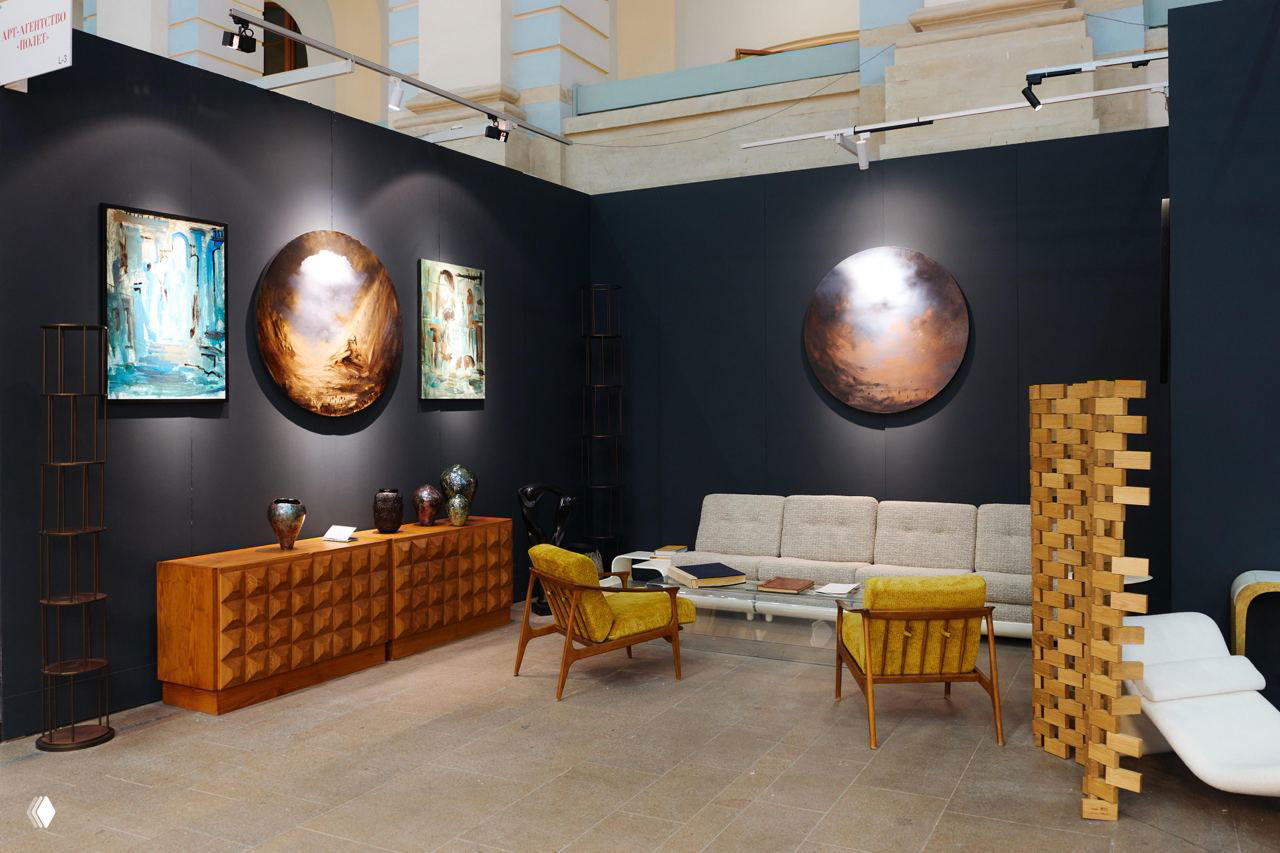 Выставочный стенд с картинами и мебелью в стиле модерн: светлый диван, деревянная консоль и круглые декоративные полотна на тёмной стене.