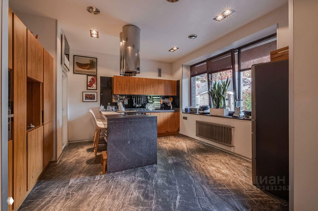 Кухня с островом и деревянными фасадами, мраморным полом, наклонной вытяжкой над рабочей зоной и большими окнами с растениями на подоконнике.