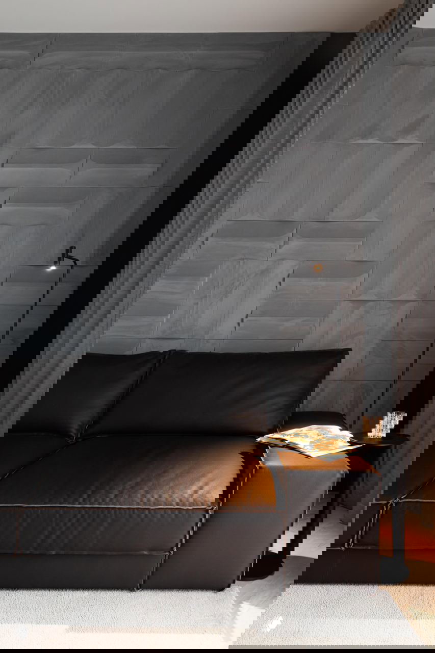 Тёмный кожаный диван на фоне рельефной графитовой панели, рядом низкий столик и направленный свет от настольной лампы для акцента в зоне.