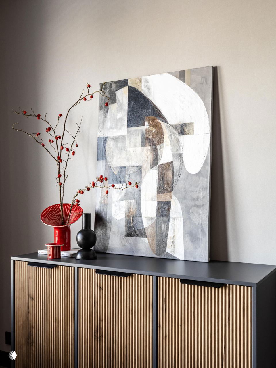 Композиция декоративных предметов: абстрактная картина, красная ваза и черные аксессуары на деревянном комоде в интерьере квартиры.