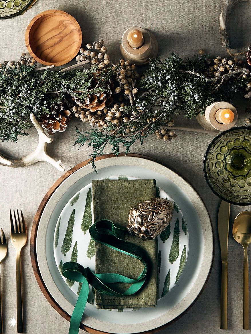 Составная сервировка в зелёных и золотых тонах: тарелки с растительным мотивом, золотая лента и натуральные шишки создают природный новогодний образ.
