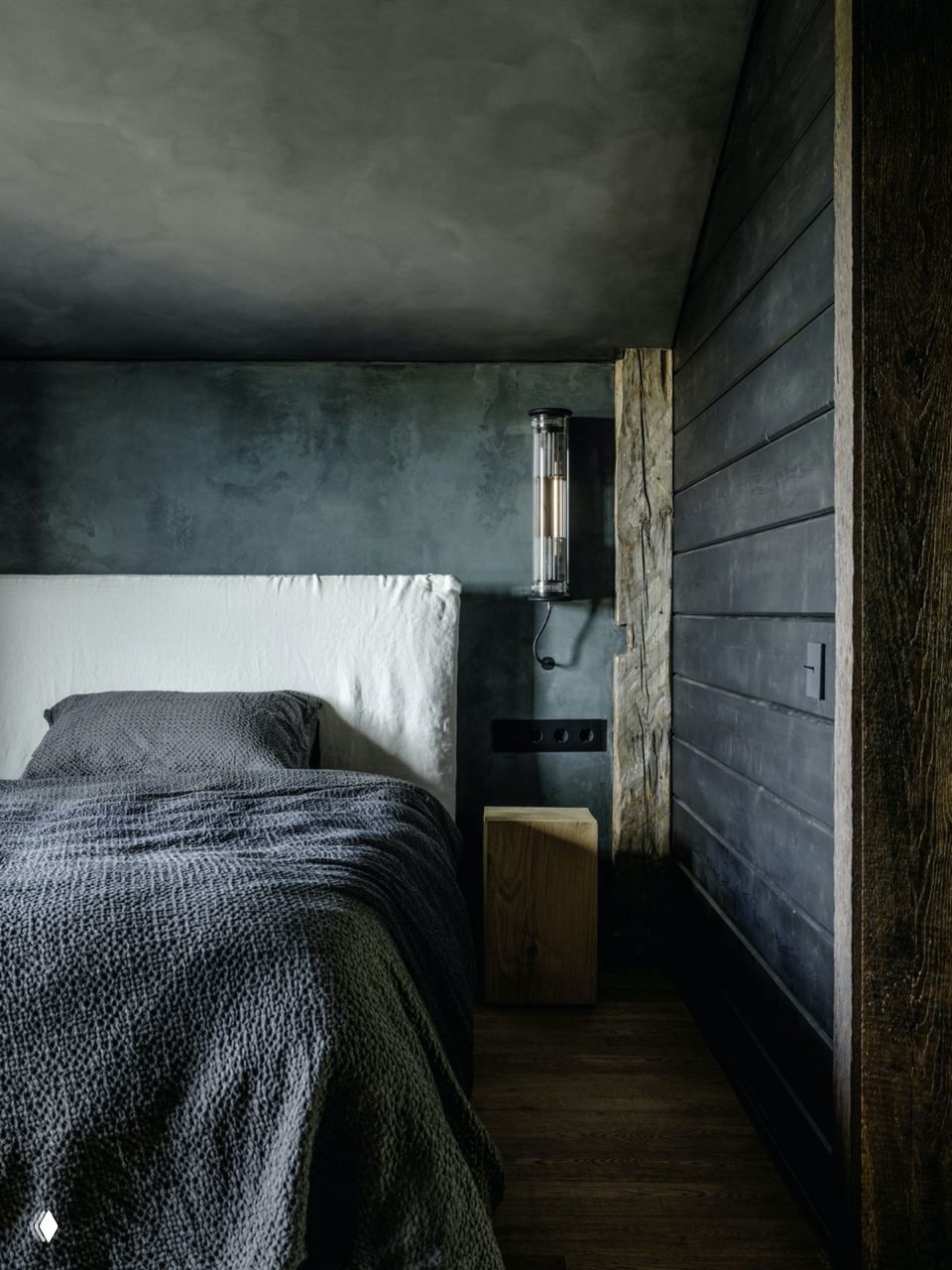 Спальня в спокойной тёмной гамме: текстильная мягкая кровать на фоне шершавых стен и деревянного пола, минимальная прикроватная тумба.