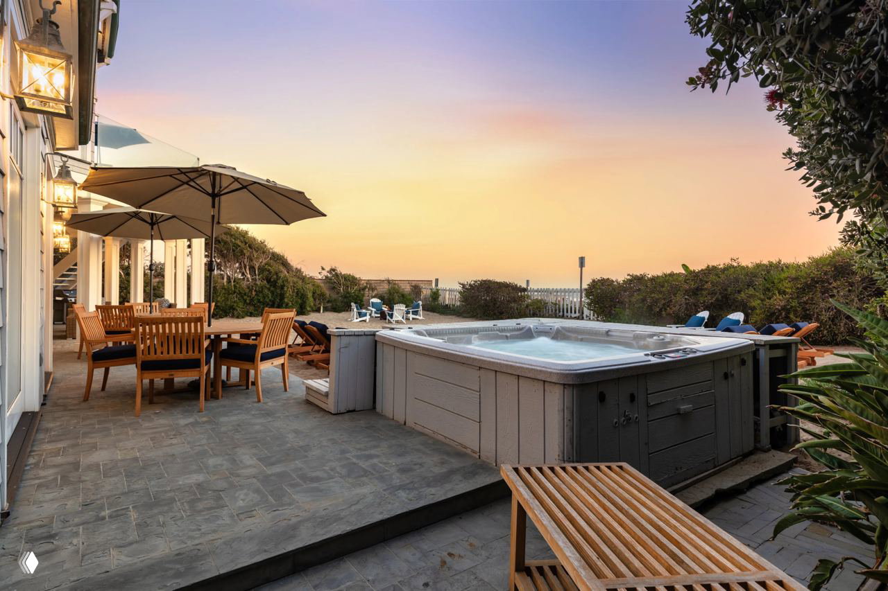 Вид на открытую террасу и внешний джакузи при закате: деревянная мебель, зонты и шезлонги у выхода к пляжу в окружении зелёных кустов.