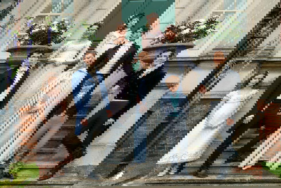 Групповой кадр актёров сериала «Бриджертоны» на ступенях усадебного дома, костюмы в стиле регентства и исторический декор вокруг входа.